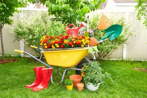 A brightly colored yellow wheelbarrow filled with vibrant red, yellow, and orange flowering plants is positioned on a lush green lawn in a garden. Surrounding the wheelbarrow are gardening tools and accessories, including a red watering can, blue and green plastic shovels, and a green watering tray. In front of the wheelbarrow, there are several small terracotta pots, some containing plants or soil, and a white plant pot with green foliage. The background features a white garden fence bordered by dense green shrubs and trees, casting dappled shade over the area. The overall scene conveys a tidy, well-maintained outdoor space suitable for lawn care and gardening activities, with natural sunlight illuminating the garden environment. This setting reflects typical gardening services offered by Lawn Mowing Forest Gate, situated within the Epping Forest area near London, ideal for enhancing outdoor spaces through professional lawn and garden maintenance.