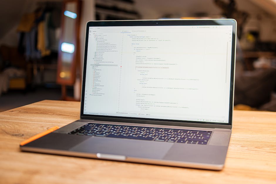 A close-up image showing a person's hand typing on a silver laptop keyboard, placed on a wooden desk. The screen displays lines of colorful code, indicating programming or website development activity
